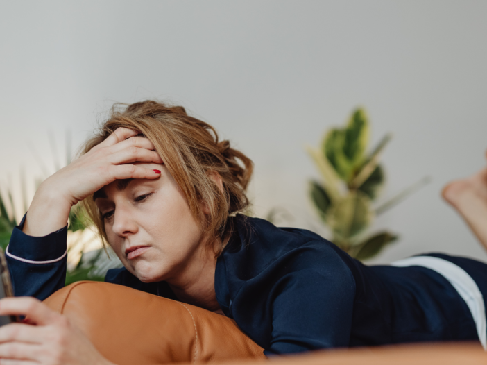 jonge vrouw ligt op de bank met mobiel in de hand en kijkt vermoeid