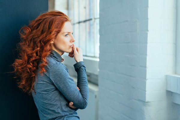 Sad lonely thoughtful young woman with gorgeous long curly red hair standing sideways indoors staring though a window|Sad lonely thoughtful young woman with gorgeous long curly red hair standing sideways indoors staring though a window