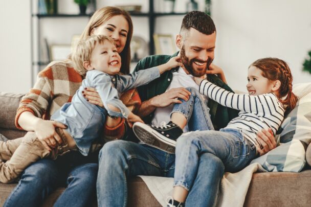 gezin van man en vrouw plus twee kinderen op de bank aan het stoeien|banner over ouderschap: foto's van gezinnen met in blauw tekstvak de tekst 'over ouderschap'