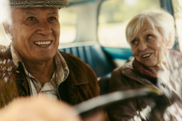 Twee ouderen mensen in een auto samen. Ze lachen heel leuk samen en de man brengt de vrouw naar haar bestemming.