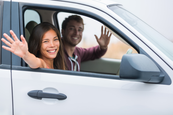 Een man en een vrouw zitten samen in een auto. De man vervoert de vrouw en ze zwaaien en lachen allebei.