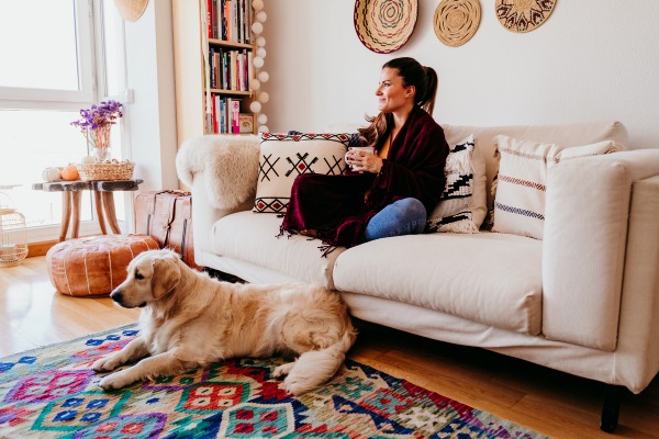 vrouw zit lekker op de bank met een kop thee. Op de grond ligt een golden retriever.