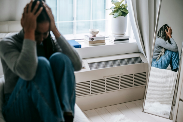 vrouw zit met haar handen in haar haar in huis. Ze maakt een verdrietige indruk.