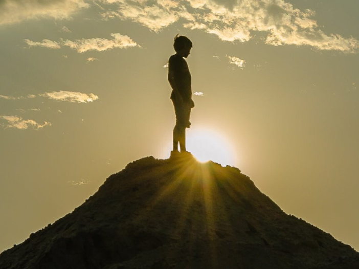 jongen staat op de top van een berg, met tegenlicht van een ondergaande zon