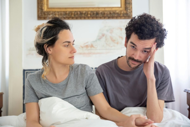 man en vrouw zitten samen rechtop in bed. Man ziet er gespannen of bezorgd uit.