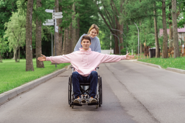 Jongen die in een rolstoel zit wordt geduwd door een meisje. Hij rijdt op de weg met armen gespreid onder de bomen.