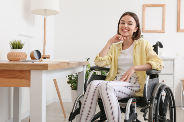 Lachende vrouw in een rolstoel. Ze heeft een geel blousje aan en ze is thuis.