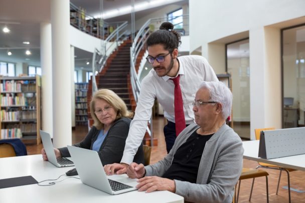 Man met een bril in een pak helpt twee oudere vrouwen met een laptop. Ze zitten in een bibliotheek.