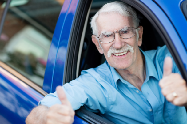 Oudere man in blauw shirt zit in een blauwe auto als passagier en steekt zijn duim op