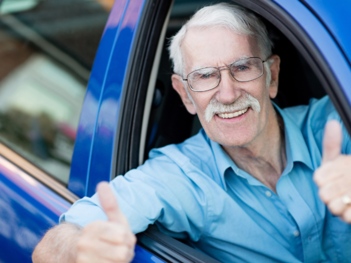 Oudere man in blauw shirt zit in een blauwe auto als passagier en steekt zijn duim op