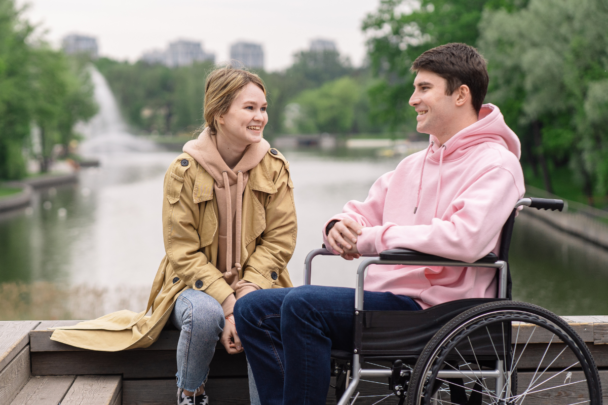 Jongen praat met meisje. Meisje met handen tussen haar benen en jongen in een rolstoel in een roze trui.