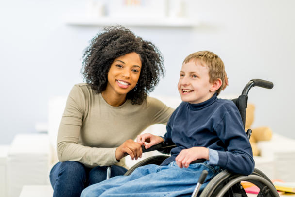 Vrouw helpt jongetje in rolstoel. De vrouw is heeft krullen en ze lachen allebei.