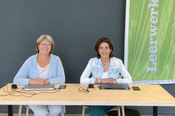 Twee vrouwen zitten naast elkaar aan een tafel. Er staat achter hun een doek met leerwerkloket erop en ze lachen.