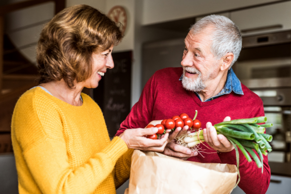 Een oudere man en een vrouw van middelbare leeftijd staan in de keuken. Ze hebben een tas met groentes vast en lachen naar elkaar.