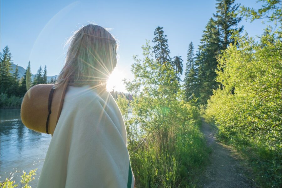 Achterkant van een vrouw met hoed die in een bosrijke omgeving loopt. De zon straalt en de lucht is blauw.