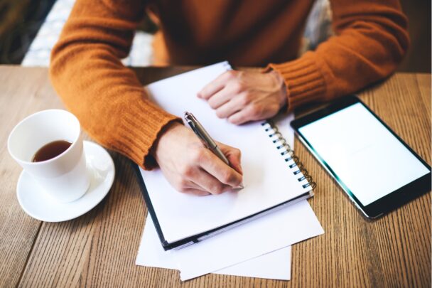 Man schrijft in een notitieboek. Er staat een kopje koffie op tafel.