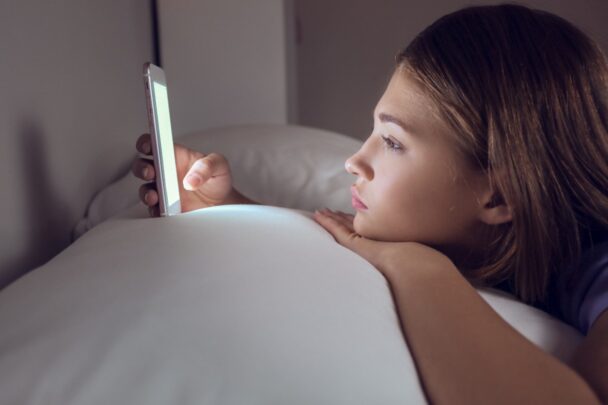 Tienermeisje ligt op bed en gebruikt haar smartphone. Ze oogt verveeld.