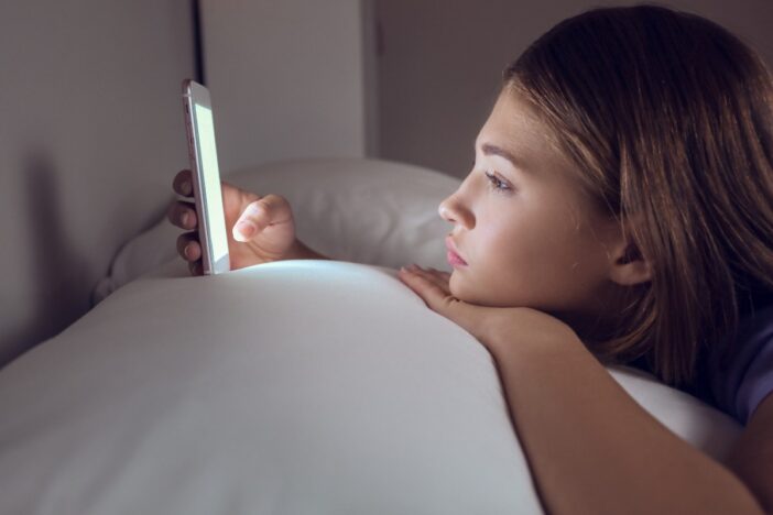 Tienermeisje ligt op bed en gebruikt haar smartphone. Ze oogt verveeld.