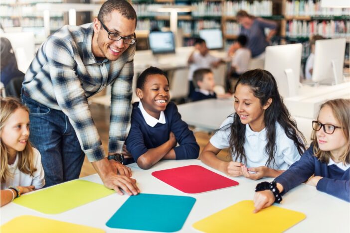 Basisschoolkinderen zitten op school aan een tafel. Op tafel ligt gekleurd papier. Er staat een volwassen man bij die uitleg geeft.