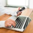 Iemand zit aan een tafel en maakt gebruik van laptop en een smartphone.