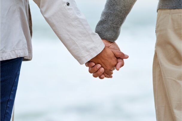 Man en vrouw houden elkaars hand vast. Op de achtergrond zien we de zee.