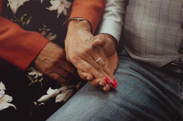 De handen van een oudere man en vrouw houden elkaar vast.