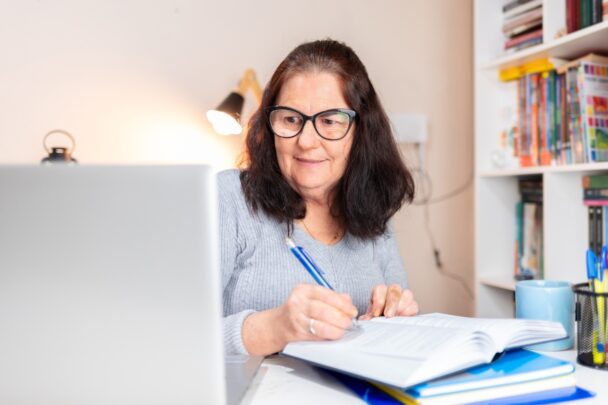 Vrouw is aan het schrijven aan een bureau met daarop boeken en een laptop.