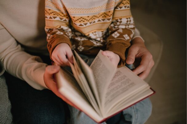 Kind zit op schoot bij volwassene en houdt een boek vast.