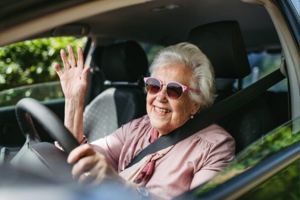 Oudere vrouw met zonnebril op rijdt in auto en zwaait naar de camera.
