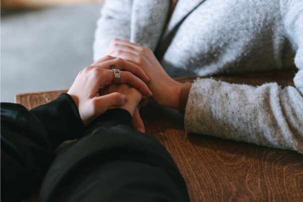 Twee vrouwen aan tafel; ze houden elkaars hand vast.