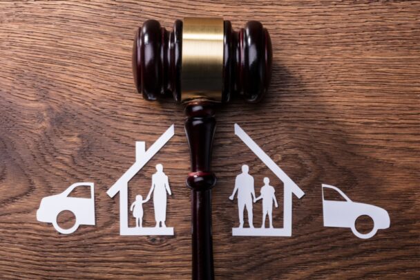 Illustratie van een rechtershamer op een houten tafel, met daarbij getekend een door de helft gedeeld huis en auto. Symboliek voor een scheiding.