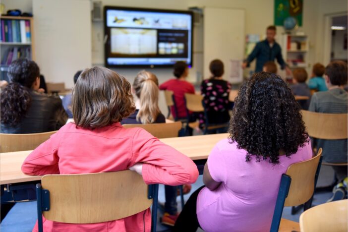 Kinderen zitten in een schoolklasje met hun rug naar de camera toe. Ze volgen een presentatie van een leraar.