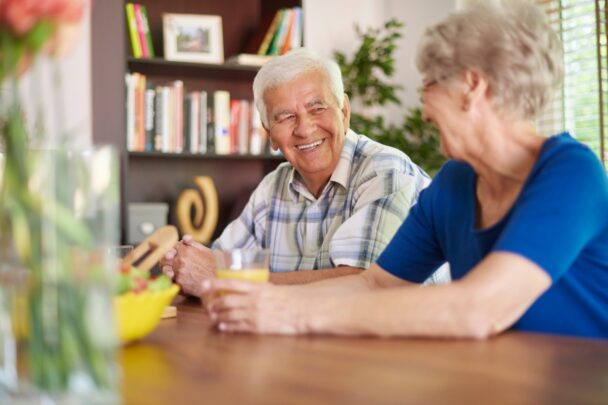 Oudere man en vrouw zitten samen aan tafel te eten en te kletsen.