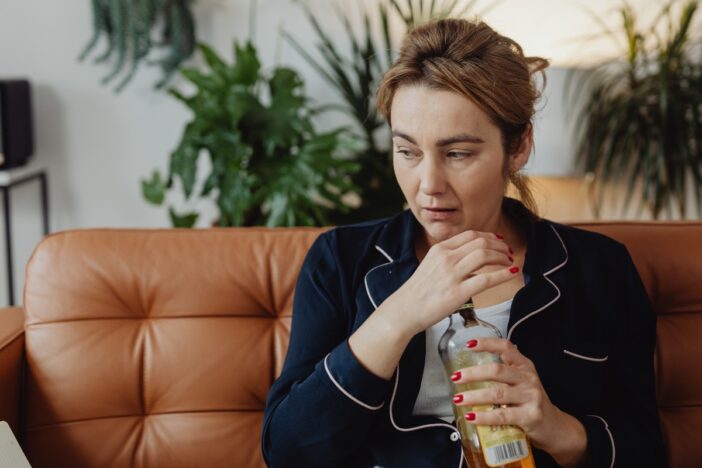 Vrouw zit op de bank met fles alcohol in haar handen. Ze oogt ongelukkig.