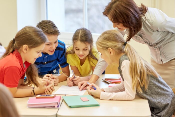 Vier kindjes werken aan een taak op papier, een schooljuf kijkt over hun schouder mee.