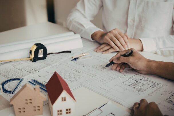 Twee mensen zitten aan tafel en bekijken papieren bouwtekeningen. Onderin twee kleine houten huisjes ter illustratie.