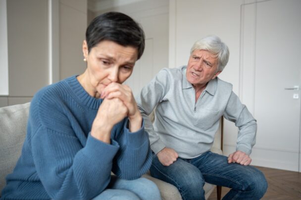 Oudere vrouw zit droevig en angstig op de bank, weggedraaid van een boze oudere man.