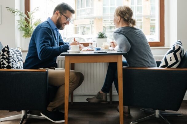 Man en vrouw voeren een gesprek aan een tafel. Achter hen is een groot raam.