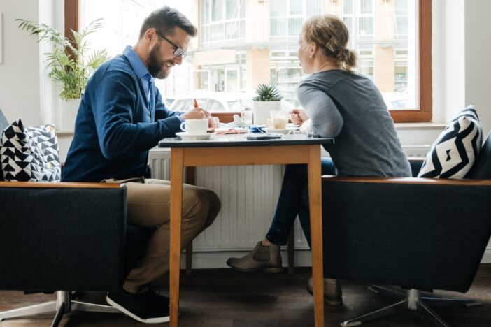 Man en vrouw voeren een gesprek aan een tafel. Achter hen is een groot raam.