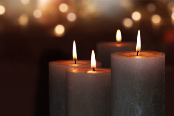 Vier brandende kaarsen tegen een donkere achtergrond met wat kleine lichtjes.