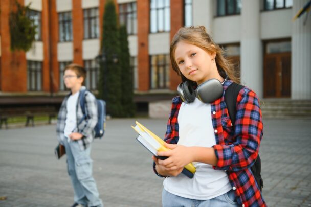 Stoere meid staat met schoolwerk en koptelefoon voor een schoolgebouw.