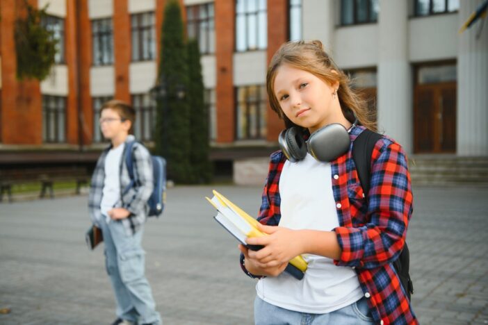 Stoere meid staat met schoolwerk en koptelefoon voor een schoolgebouw.